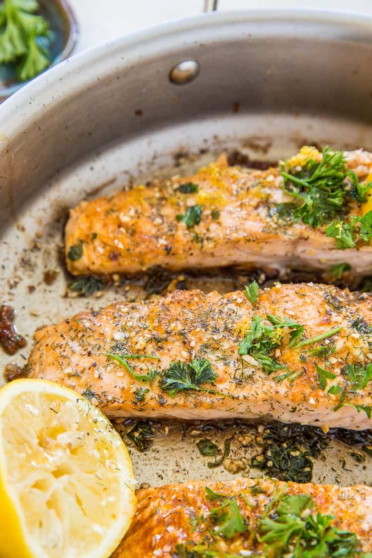 Two pan-fried salmon fillets in a metal pan, garnished with chopped herbs and spices, with a halved lemon placed beside the fillets.