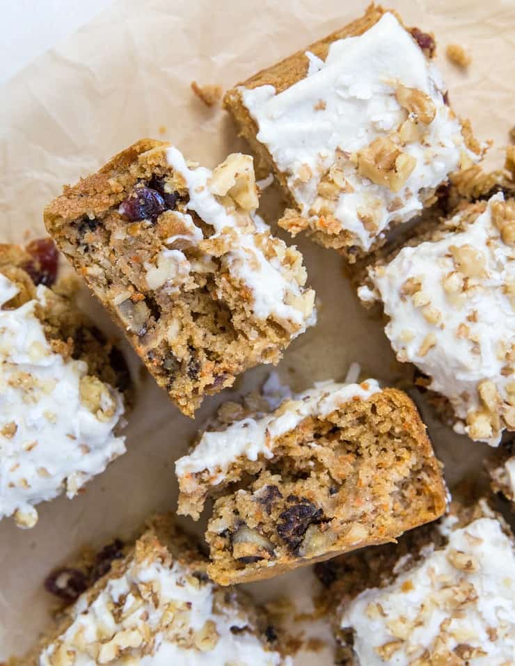 Sliced pieces of low-carb carrot cake topped with white frosting and chopped walnuts, showing a moist, crumbly texture with visible bits of carrot and raisins, arranged on parchment paper.