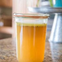 A glass jar filled with golden-yellow Instant Pot Chicken Bone Broth sits on a kitchen countertop, a layer of fat floating on top. The background is slightly blurred, showing kitchen elements and a potted plant.