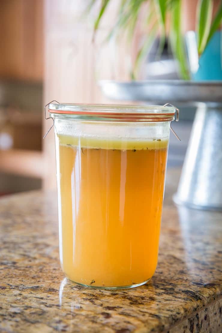 A glass jar filled with golden-yellow Instant Pot Chicken Bone Broth sits on a kitchen countertop, a layer of fat floating on top. The background is slightly blurred, showing kitchen elements and a potted plant.
