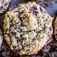 A close-up of a freshly baked keto chocolate chip cookie with a rough texture, golden brown edges, and visible chunks of melted chocolate on a baking sheet.