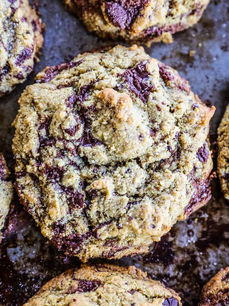 A close-up of a freshly baked keto chocolate chip cookie with a rough texture, golden brown edges, and visible chunks of melted chocolate on a baking sheet.