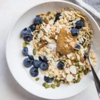 A bowl of cereal topped with fresh blueberries, sliced almonds, pumpkin seeds, and a dollop of nut butter sits beside a spoon and striped cloth, featuring creamy Instant Pot Coconut Milk Yogurt for a delicious touch.