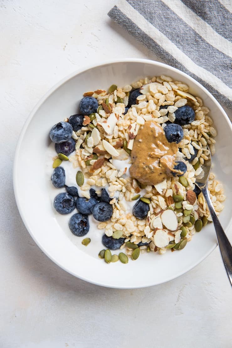 A bowl of cereal topped with fresh blueberries, sliced almonds, pumpkin seeds, and a dollop of nut butter sits beside a spoon and striped cloth, featuring creamy Instant Pot Coconut Milk Yogurt for a delicious touch.