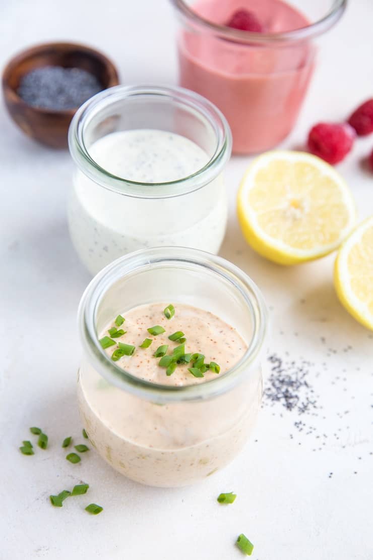 Three glass jars with different creamy keto salad dressings, garnished with chives, poppy seeds, and raspberries. Lemon halves, chives, poppy seeds, and raspberries are arranged around the jars on a light surface.