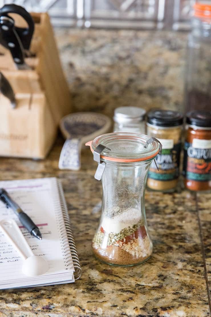 Jar with a spice mix inside to make taco seasoning, and a spiral notebook next to the jar with the recipe and jars of seasoning in the background.