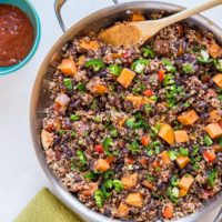 A Mexican vegan skillet filled with quinoa, black beans, diced sweet potatoes, chopped tomatoes, and sliced jalapeños is shown with a wooden spoon inside. A bowl of red salsa and a green cloth napkin sit beside the pan.