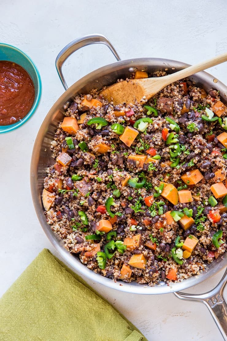 A Mexican vegan skillet filled with quinoa, black beans, diced sweet potatoes, chopped tomatoes, and sliced jalapeños is shown with a wooden spoon inside. A bowl of red salsa and a green cloth napkin sit beside the pan.