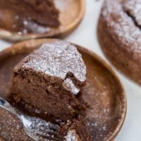 A slice of gluten-free chocolate water cake dusted with powdered sugar sits on a wooden plate, with a fork holding a bite-sized piece in front. More cake slices and a whole cake are visible in the background.