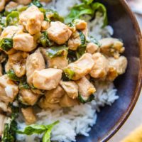 A bowl of white rice topped with Thai Basil Chicken—tender stir-fried chicken pieces and fresh green basil leaves—served in a dark ceramic bowl.