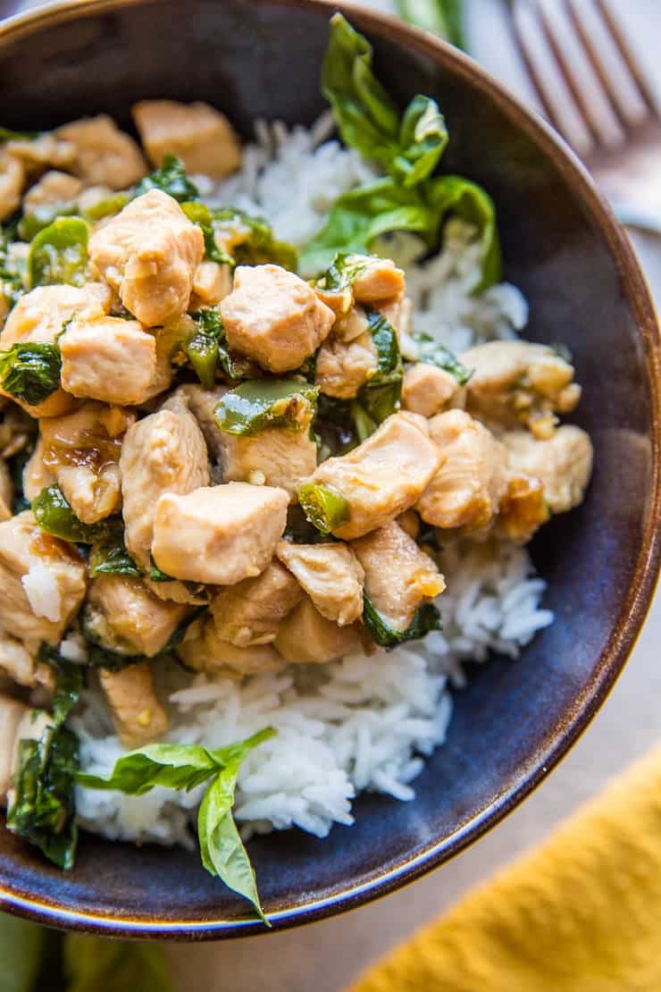 A bowl of white rice topped with Thai Basil Chicken—tender stir-fried chicken pieces and fresh green basil leaves—served in a dark ceramic bowl.