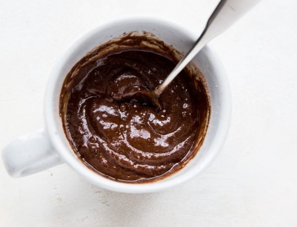 A white mug filled with thick, single serve brownie batter, with a metal spoon sticking out, sits on a light surface.