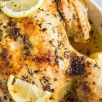 A close-up of a garlic herb rotisserie chicken garnished with fresh herbs and lemon slices, served on a white plate. The chicken boasts golden-brown, crispy skin and visible seasoning.