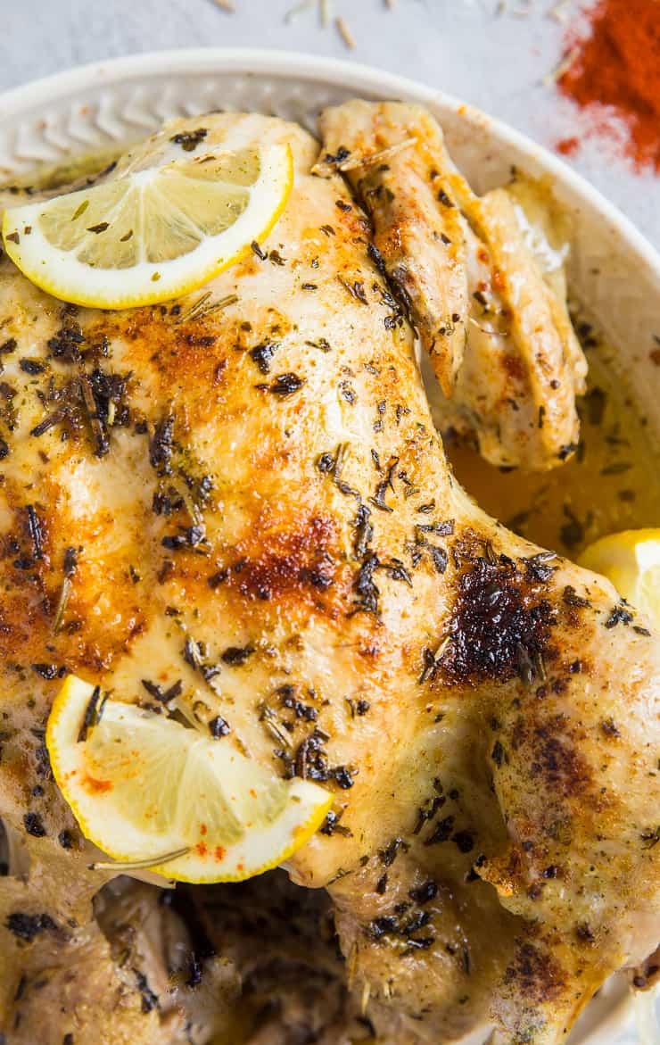 A close-up of a garlic herb rotisserie chicken garnished with fresh herbs and lemon slices, served on a white plate. The chicken boasts golden-brown, crispy skin and visible seasoning.
