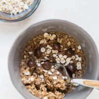 A gray bowl filled with oatmeal, chocolate chips, and a spoon evokes the cozy feel of an oatmeal cookie in a mug. Next to it is a small bowl of dry oats, all set on a light-colored surface.