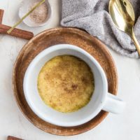 A white mug filled with a Keto Snickerdoodle Mug Cookie, topped with cinnamon, sits in a wooden bowl. Nearby are a golden spoon, gray cloth napkin, small dish of cinnamon sugar, and cinnamon sticks on a light surface.