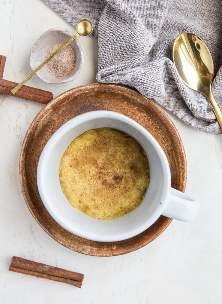 A white mug filled with a Keto Snickerdoodle Mug Cookie, topped with cinnamon, sits in a wooden bowl. Nearby are a golden spoon, gray cloth napkin, small dish of cinnamon sugar, and cinnamon sticks on a light surface.