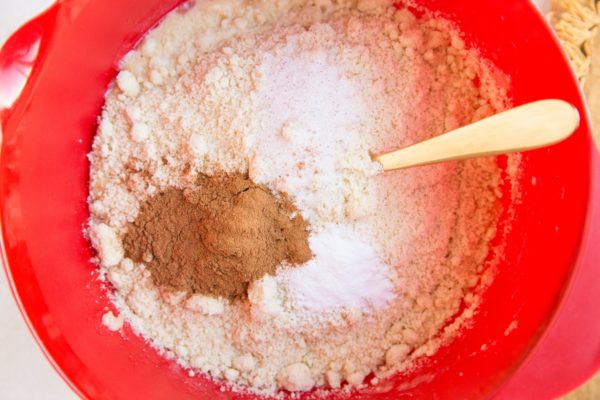A red mixing bowl containing flour, baking powder, cinnamon, and salt with a wooden spoon resting inside—perfect for making Healthy Carrot Cake Muffins or even Paleo Carrot Cake Muffins.
