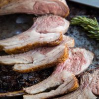 Sliced rack of lamb with a crispy seasoned crust and juicy pink center, arranged on a rustic baking sheet next to a sprig of rosemary and a carving knife—perfect inspiration if you’re wondering how to cook lamb chops at home.