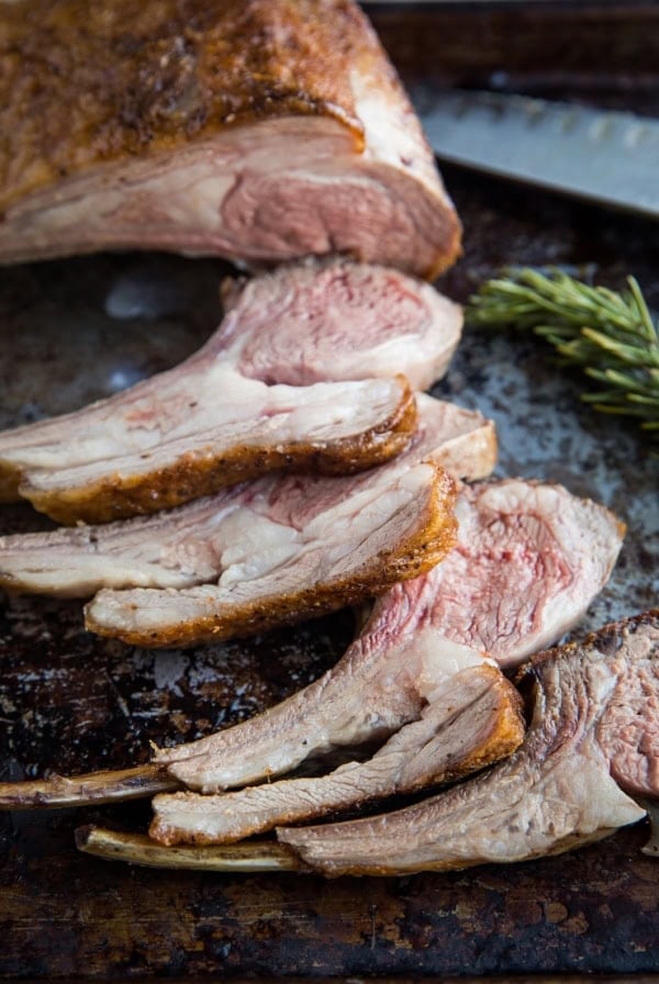 Sliced rack of lamb with a crispy seasoned crust and juicy pink center, arranged on a rustic baking sheet next to a sprig of rosemary and a carving knife—perfect inspiration if you’re wondering how to cook lamb chops at home.