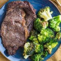 Blue plate with steak and broccoli on it with a gold fork to the side and a golden napkin