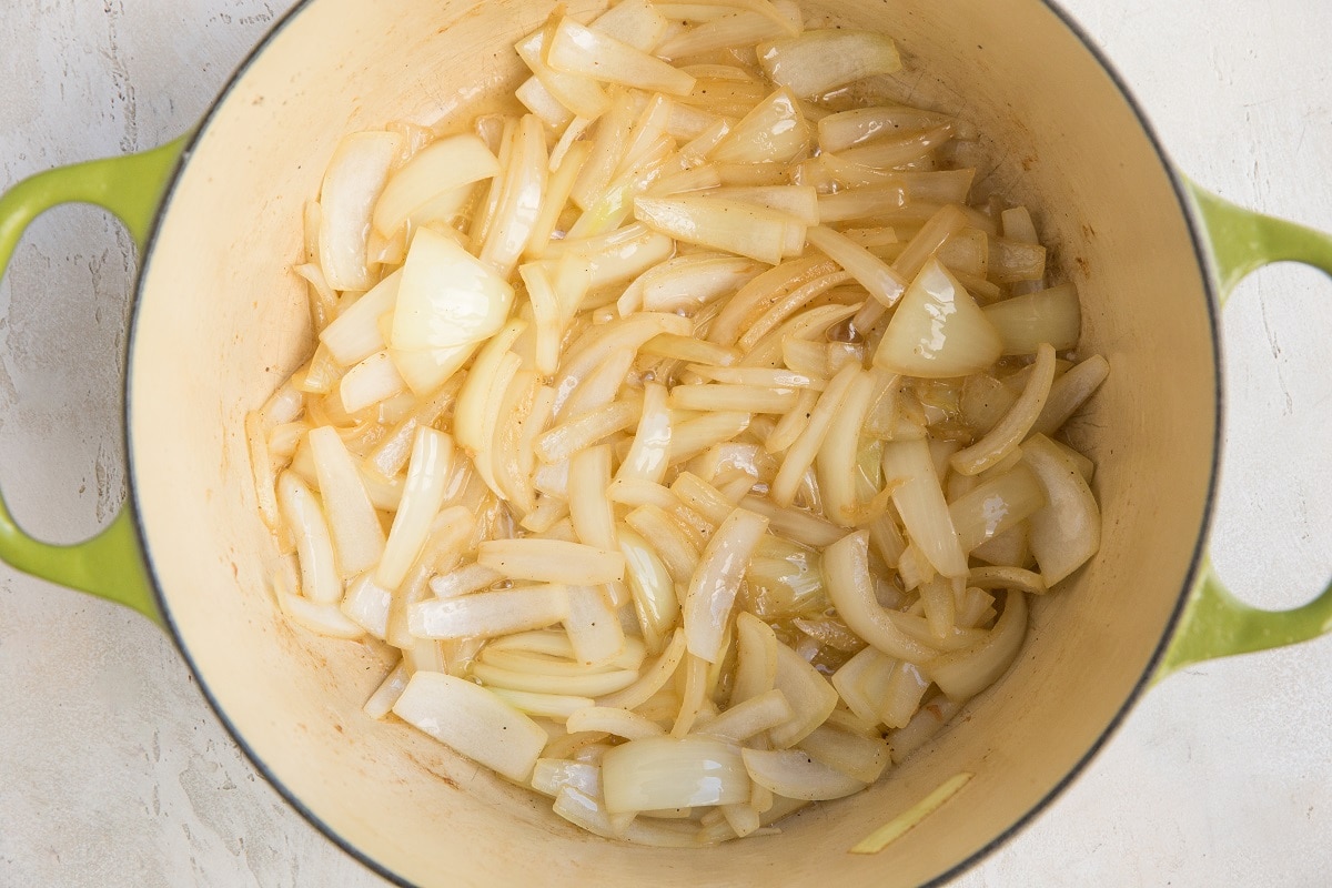 A green Dutch oven filled with sliced onions being sautéed for Chicken Cacciatore, starting to turn translucent. The pot sits on a light-colored surface.