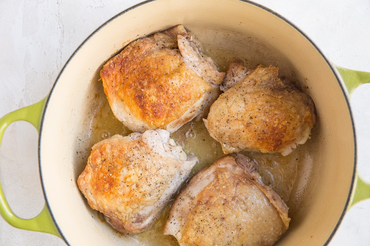 Four browned and seasoned chicken thighs cooking in a light green Dutch oven with some oil at the bottom, perfect as the base for a flavorful Chicken Cacciatore.