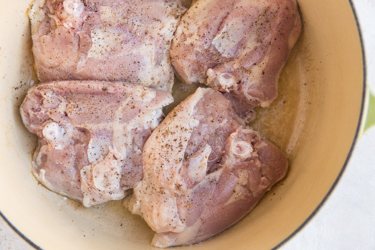 Four raw chicken thighs seasoned with salt and pepper are placed in a light-colored pot, ready to be cooked for a savory Chicken Cacciatore.