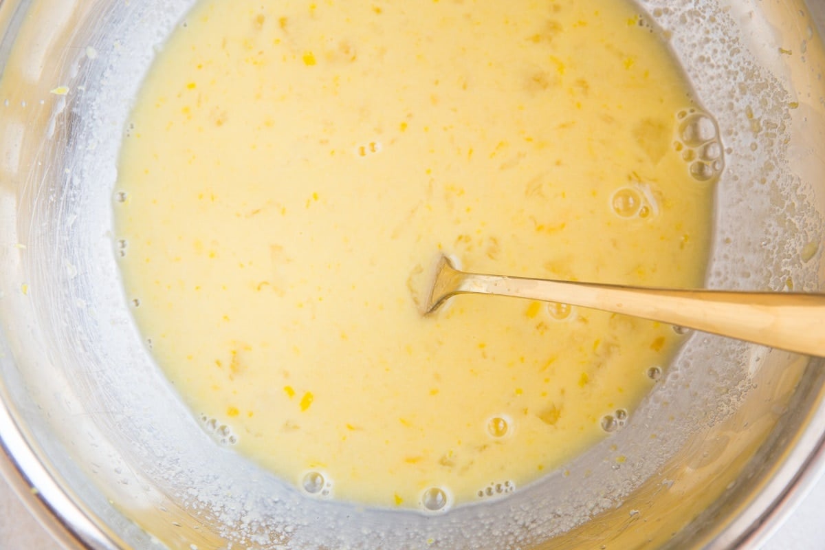 A metal mixing bowl filled with pale yellow batter, flecked with small bits for Keto Lemon Bars, with a metal spoon resting inside.