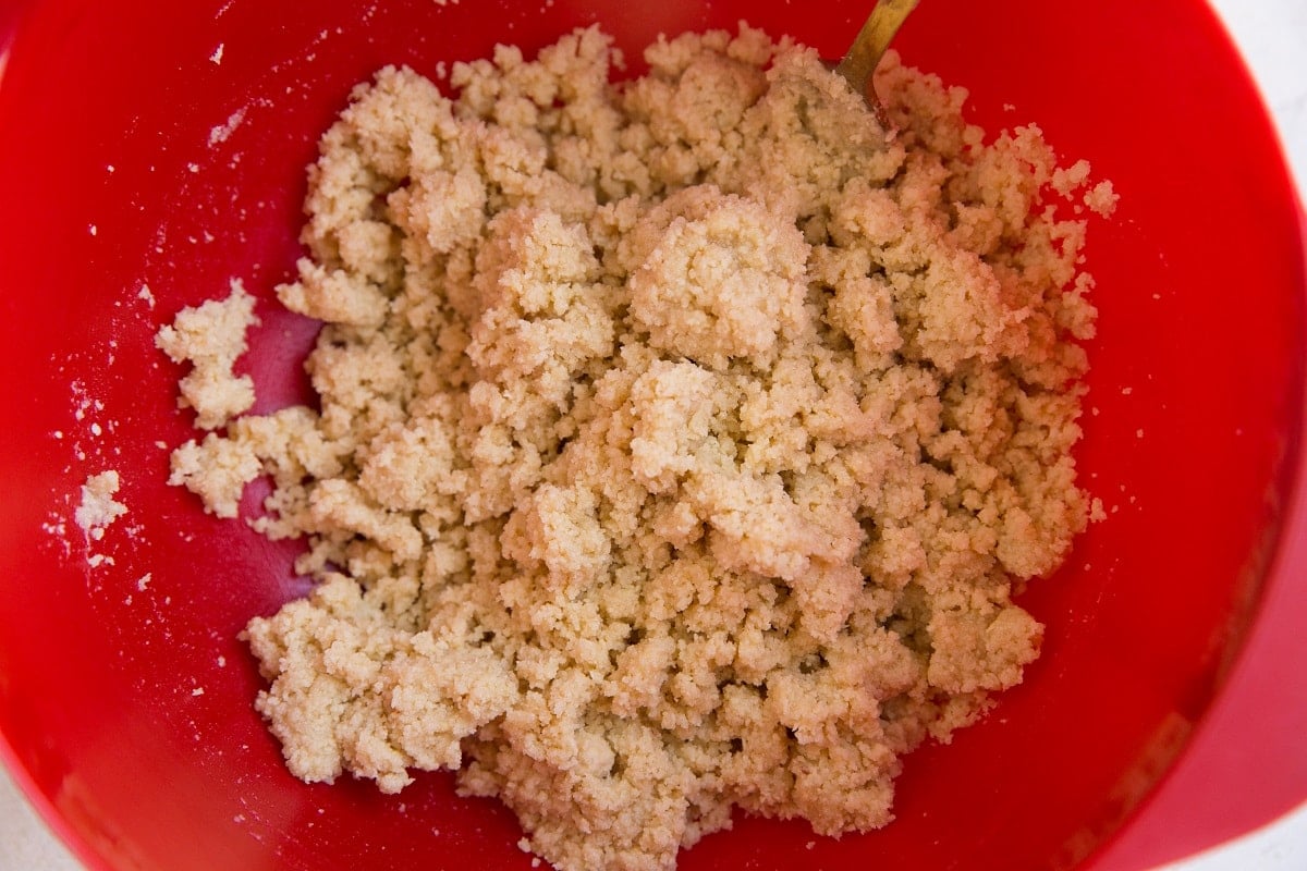 A red mixing bowl filled with crumbly, light brown Keto Lemon Bars dough mixture, with part of a metal spoon visible in the upper right corner.