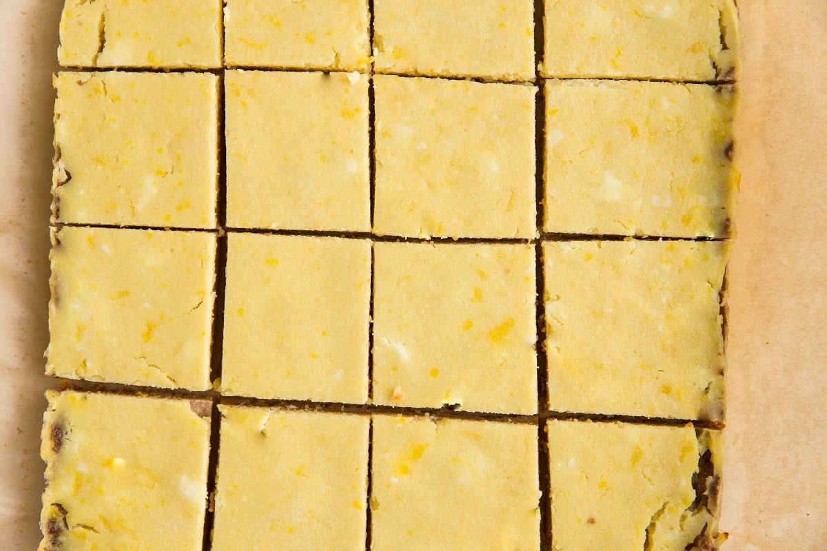 A close-up of a tray of Keto Lemon Bars cut into even squares shows a yellow, slightly textured top layer with tiny specks, all arranged on parchment paper.