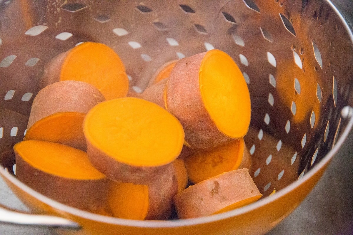 Steaming orange sweet potato slices with skin on, perfect for making smashed sweet potatoes, rest in a metal colander with holes. Wisps of steam rise from the warm, tender sweet potatoes.