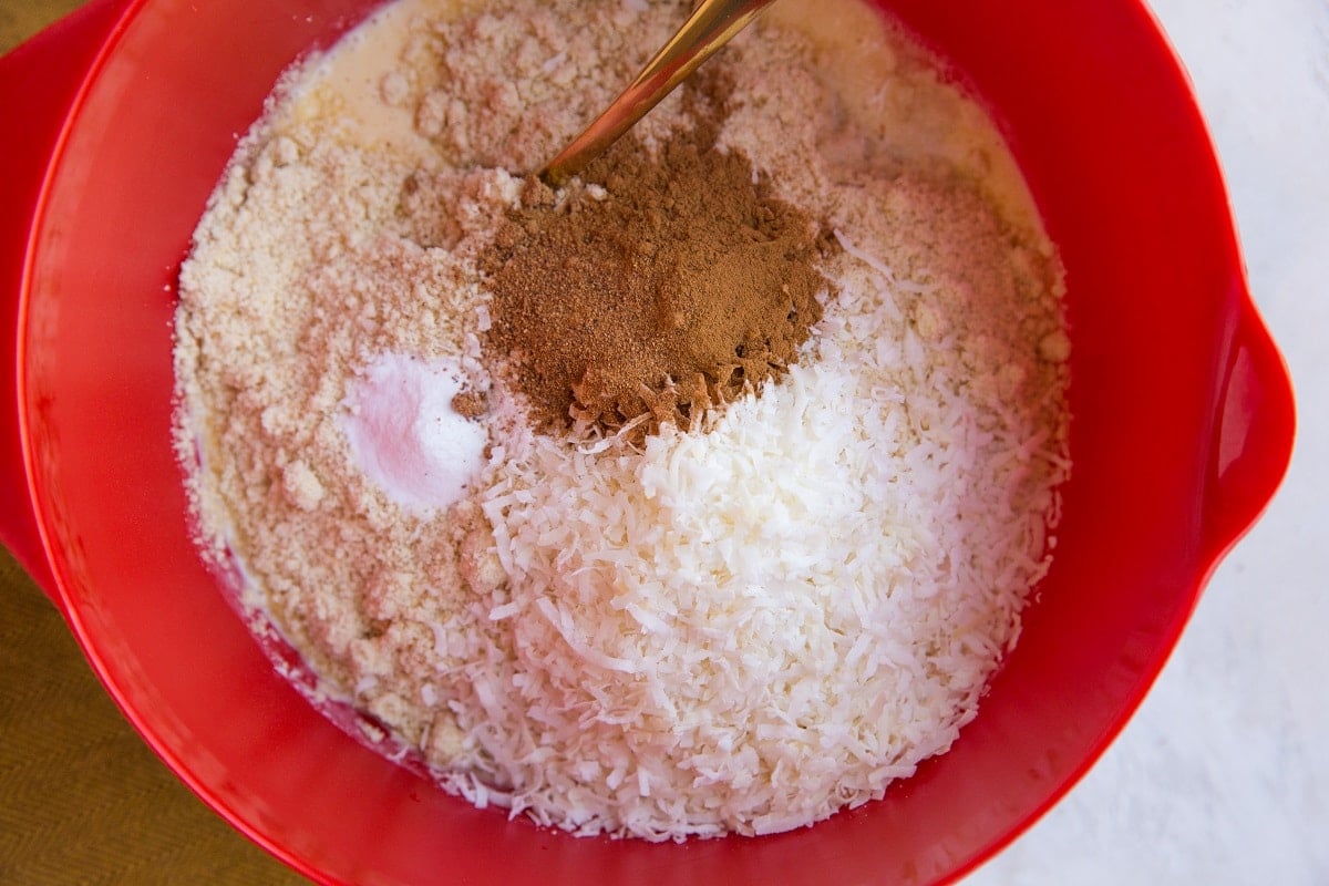 A red mixing bowl filled with shredded coconut, almond flour, cinnamon, baking powder, and other dry ingredients—perfect for making keto carrot cake muffins—with a spoon partially visible in the mixture.