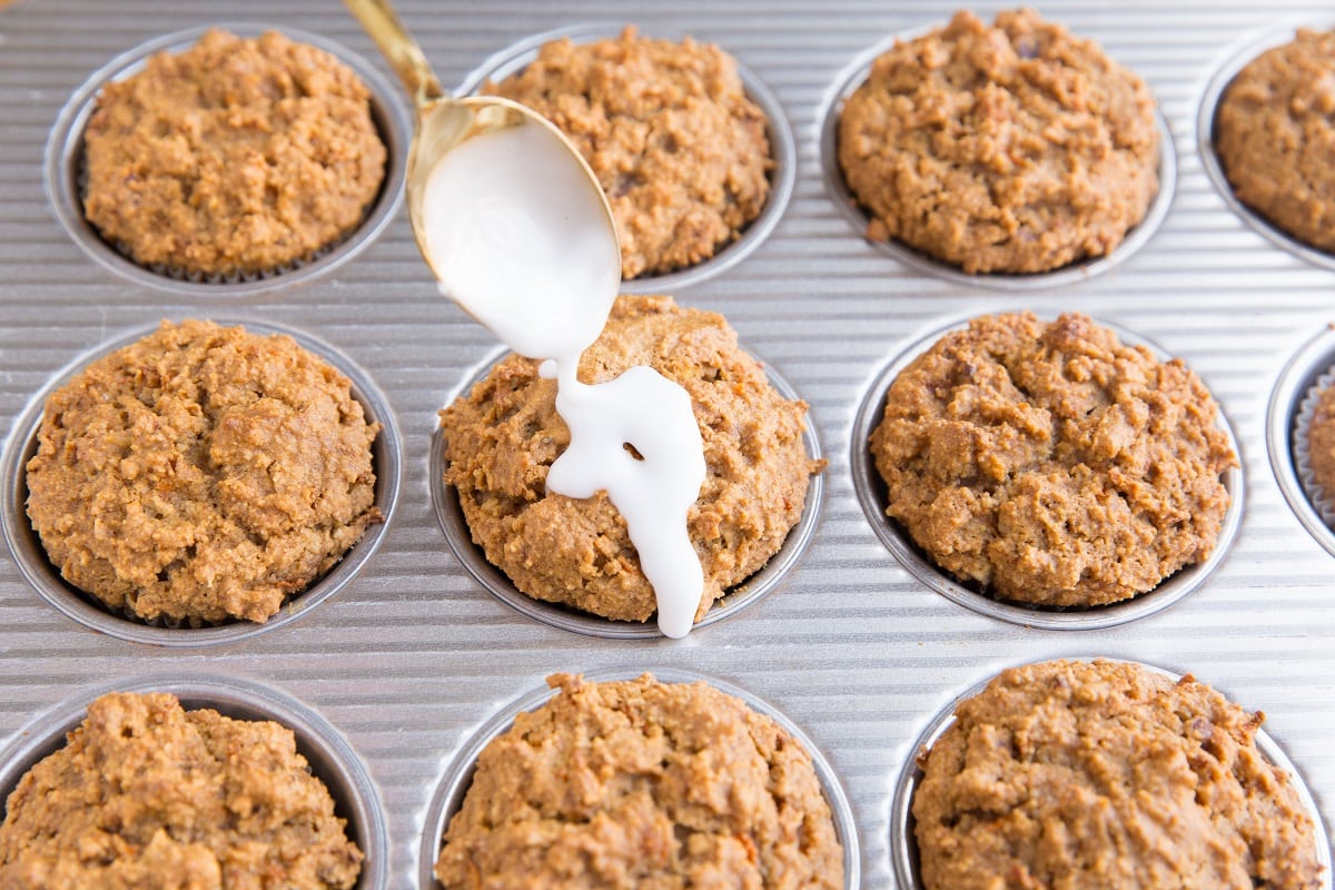 A spoon drizzles white glaze over a batch of freshly baked keto carrot cake cupcakes in a muffin tin.