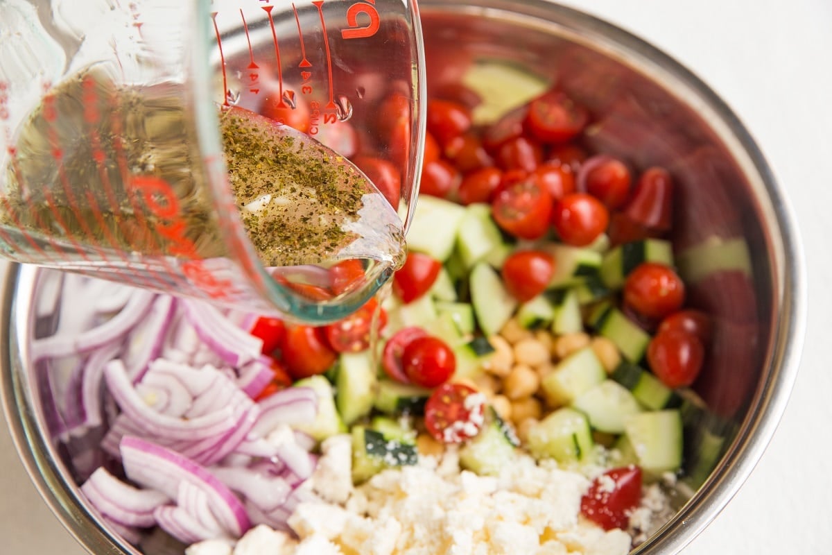 A glass measuring cup pours vinaigrette dressing over a bowl filled with chopped cucumbers, cherry tomatoes, red onion slices, chickpeas, and crumbled feta cheese for a fresh Greek chickpea salad.