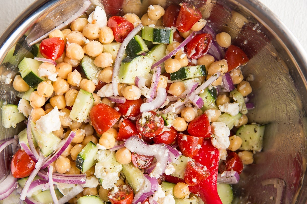 A close-up of a bowl filled with a vibrant Mediterranean chickpea salad, featuring sliced cucumbers, cherry tomatoes, red onion, crumbled feta cheese, fresh herbs, and hearty chickpeas.