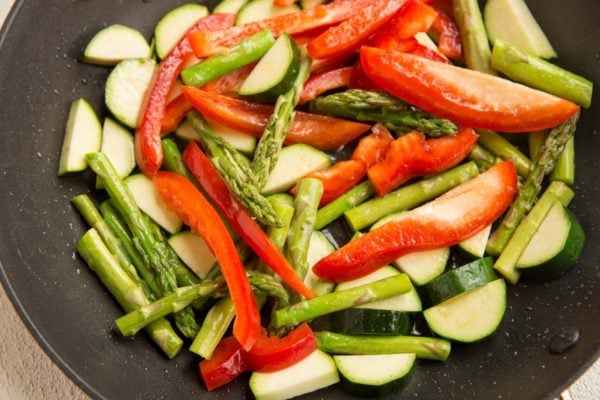 Sliced red bell peppers, zucchini, and asparagus spears being sautéed in a black skillet for a fresh pasta primavera recipe.
