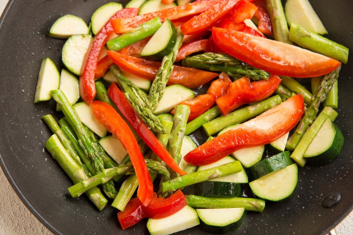 Sliced red bell peppers, zucchini, and asparagus spears being sautéed in a black skillet for a fresh pasta primavera recipe.