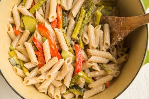A pot of creamy pasta primavera with penne, sliced red bell peppers, zucchini, and asparagus is being stirred with a wooden spoon.