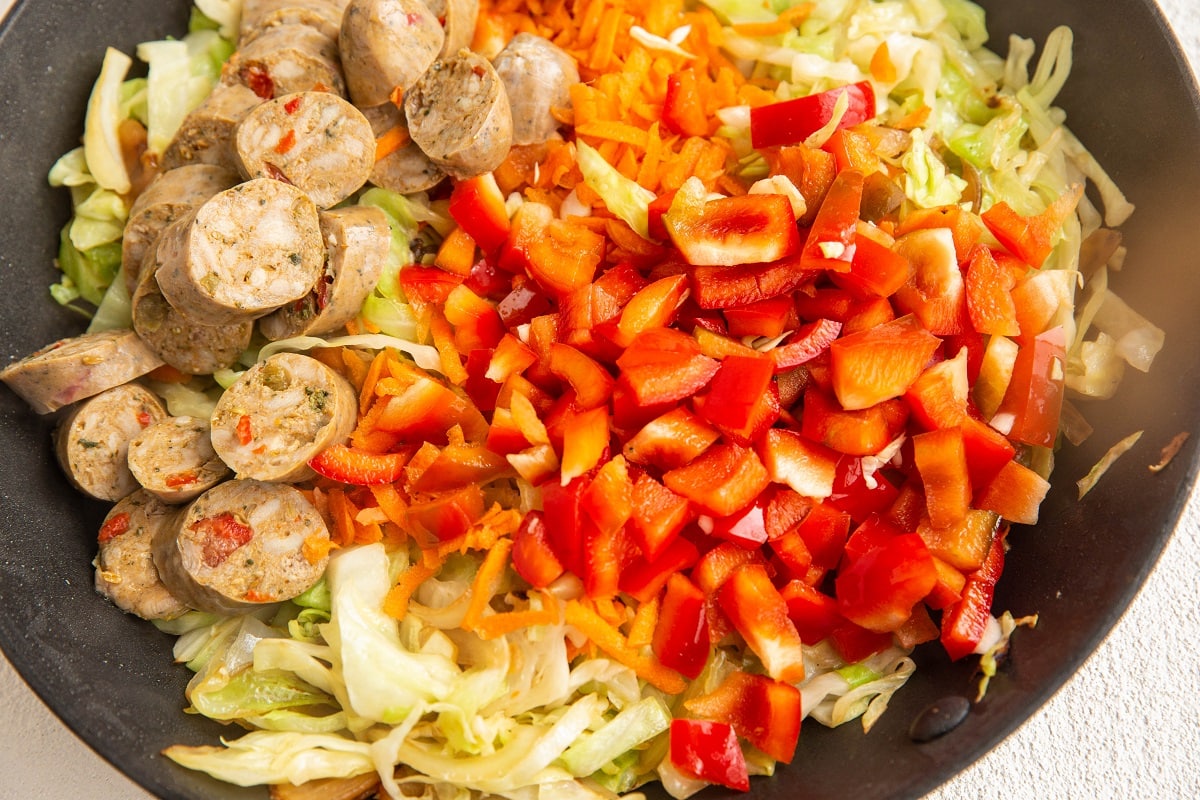 Sliced sausage, chopped red bell pepper, shredded carrots, and cabbage cooking together in a skillet for an easy sausage and cabbage recipe.