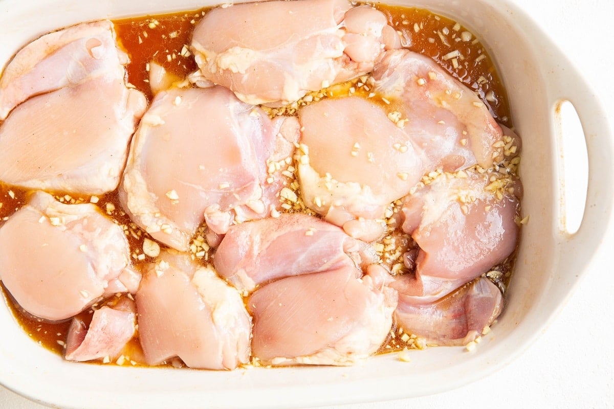 Raw chicken thighs marinating in a white dish with soy sauce, minced garlic, and other seasonings—perfect for honey garlic baked chicken thighs. The chicken is partially submerged in the flavorful marinade.