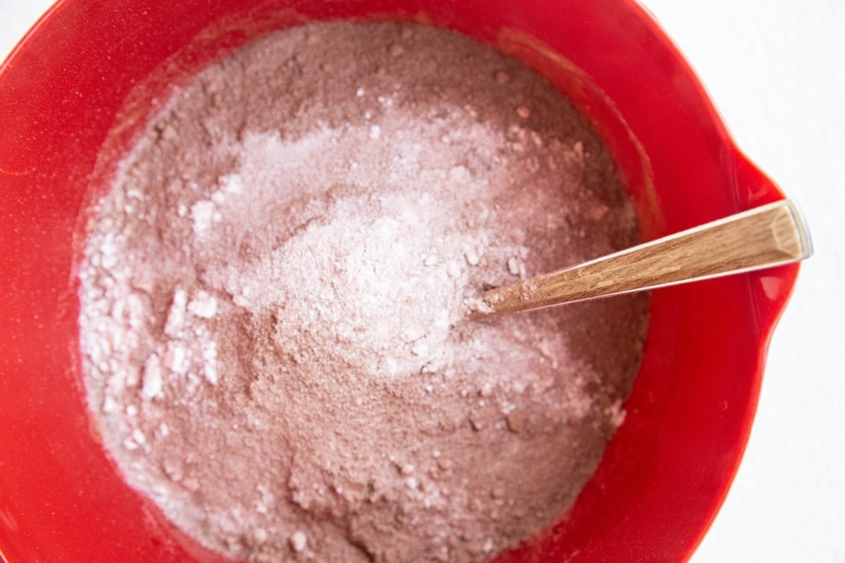 A red mixing bowl filled with dry coconut flour brownie mix powder, with a wooden spoon resting inside the bowl.