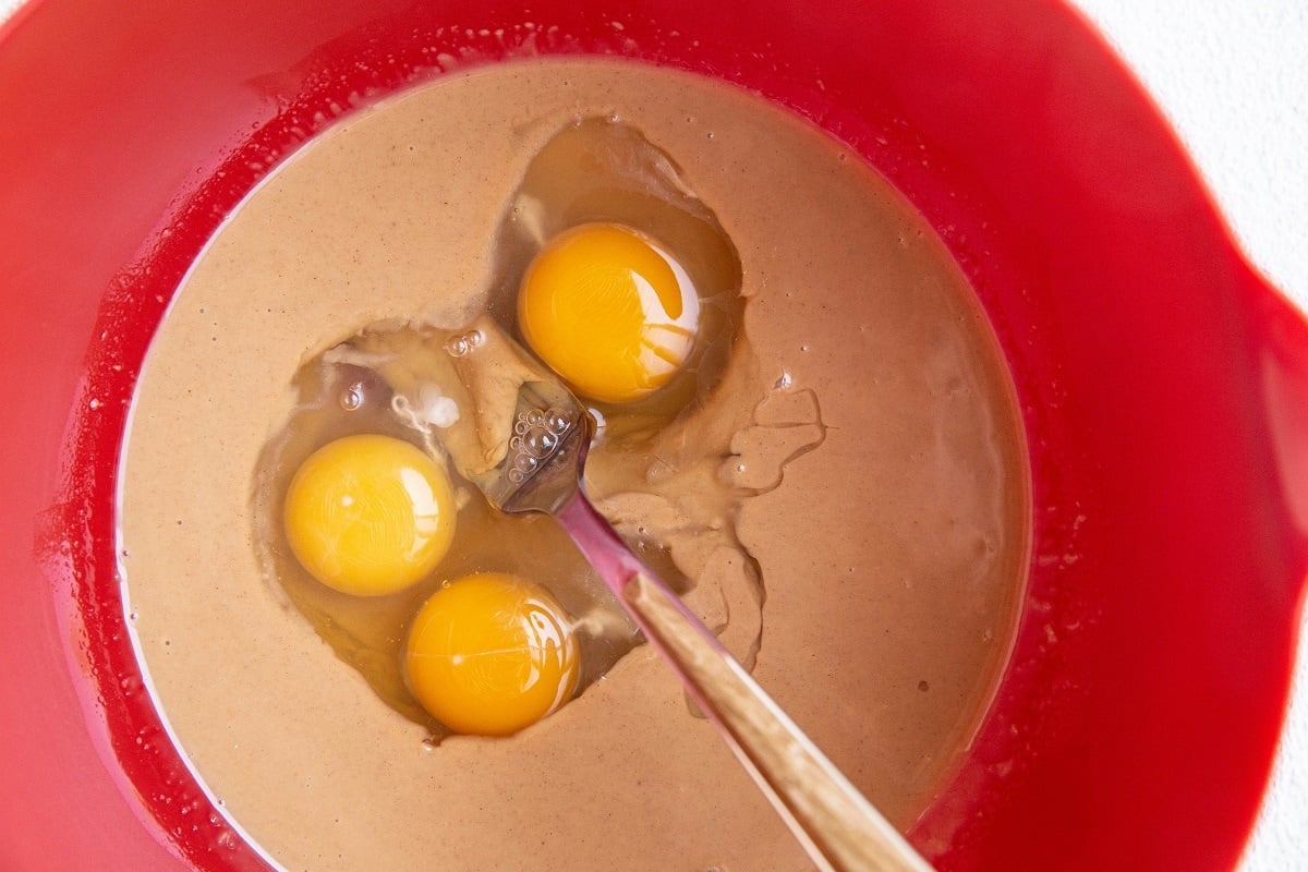 A red mixing bowl containing a light brown batter for keto coconut flour brownies, with three raw eggs cracked on top and a metal fork partially submerged in the mixture.