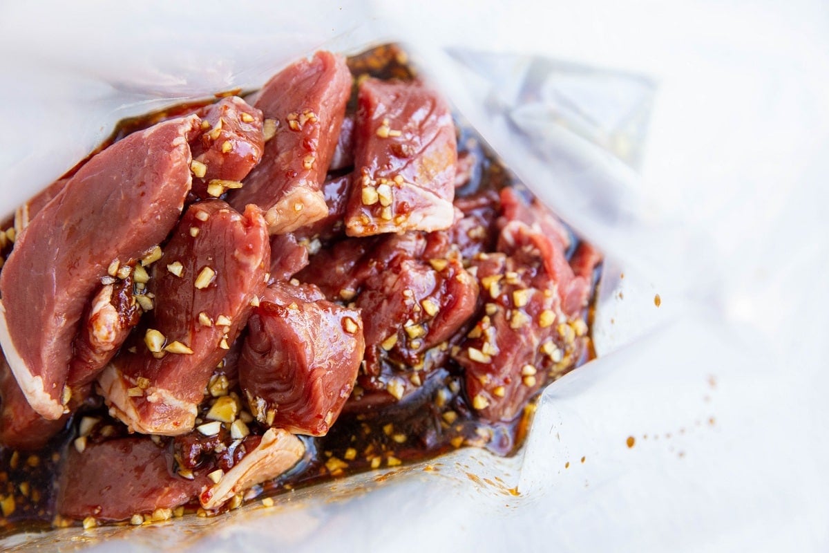Raw pieces of beef marinating in a clear plastic bag with a mixture of soy sauce, chopped garlic, and spices—classic flavors for Beef Bulgogi—coat the meat, giving it a glossy appearance.