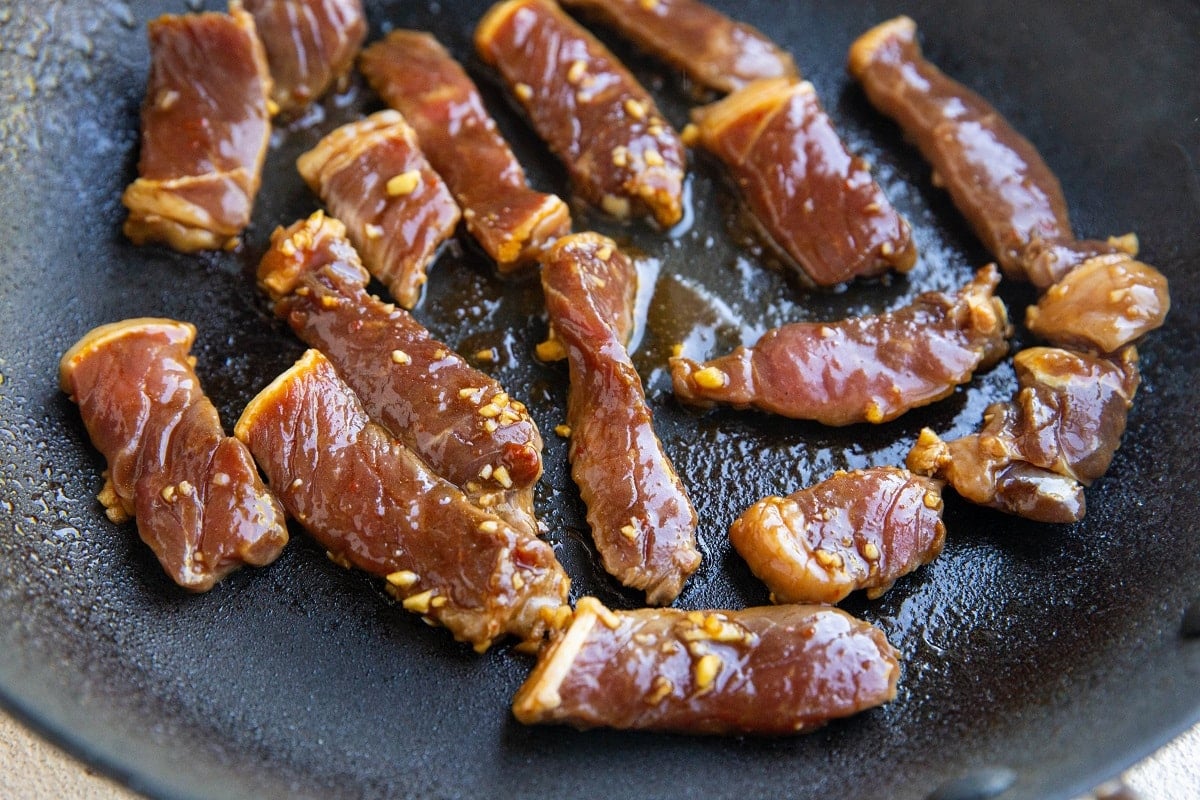 Strips of marinated bulgogi beef with visible bits of garlic are cooking in a black skillet, starting to brown and sizzle in oil.