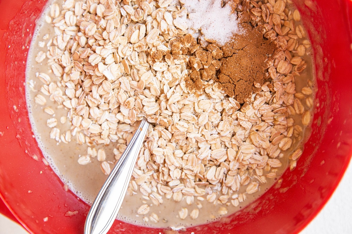 A close-up of a red mixing bowl containing the base for oatmeal muffins—rolled oats, brown spices, salt, and liquid—with a silver spoon partially submerged in the mixture. Perfect for Peanut Butter Banana Baked Oatmeal Muffins.