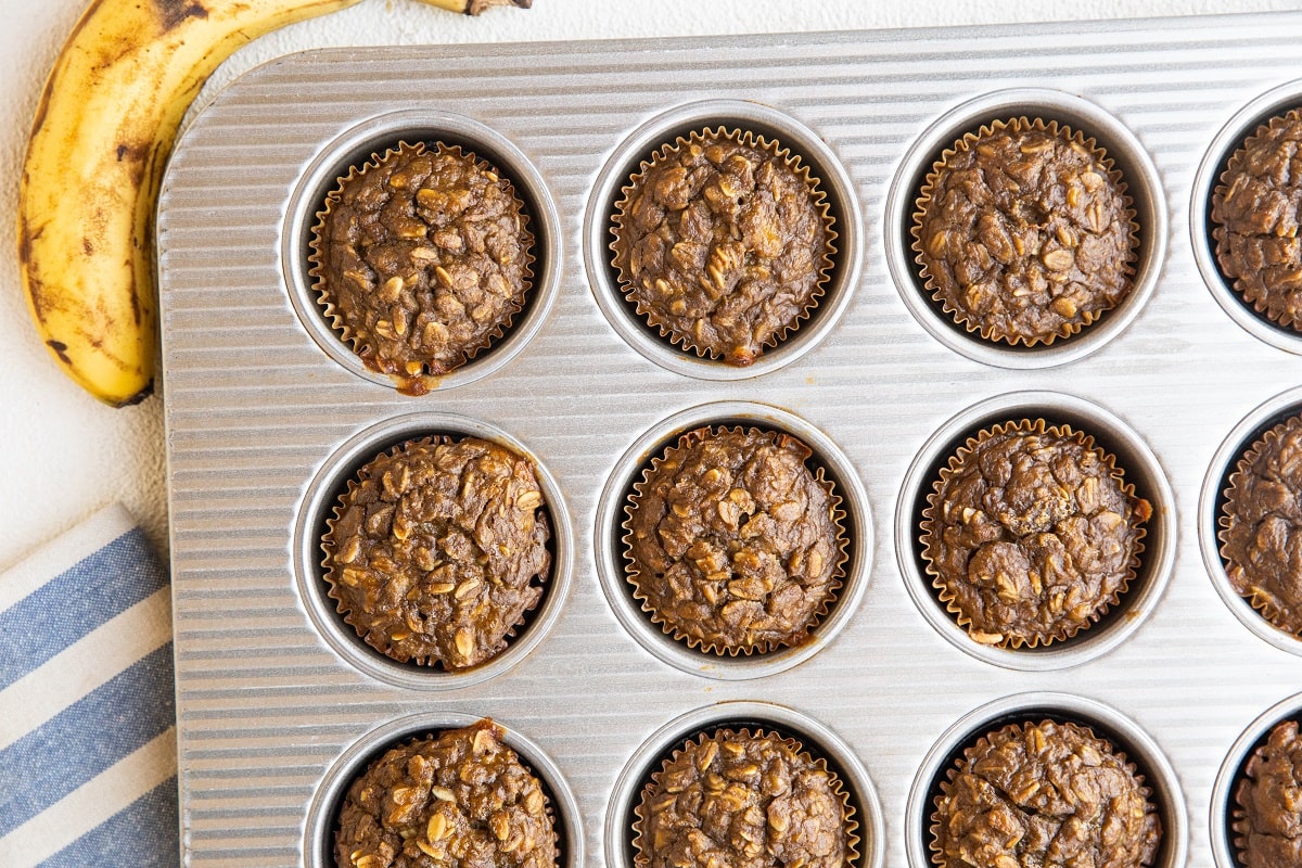 A muffin tin filled with twelve baked chocolate banana oatmeal muffins, perfect for Peanut Butter Banana Baked Oatmeal Muffins fans, with a ripe banana and a blue-striped cloth visible beside it.