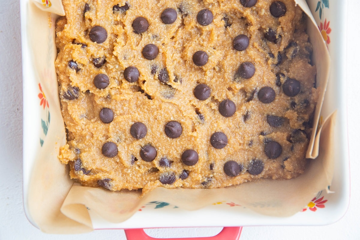 An 8-inch square baking pan lined with parchment paper with an even layer of pumpkin blondie dough, sprinkled with chocolate chips.