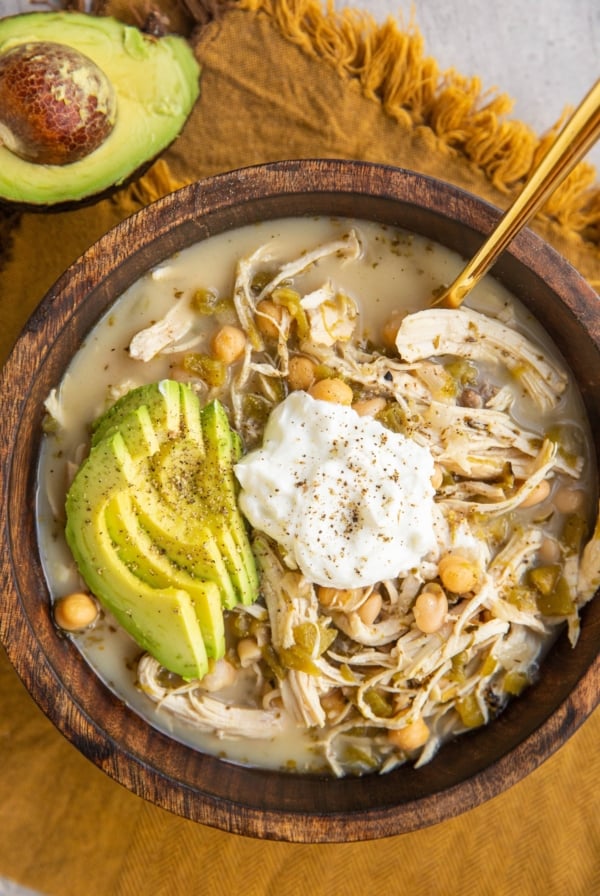 Big bowl of chili with Greek yogurt on top, sliced avocado and a gold spoon in the soup. A golden napkin underneath the bowl and half of an avocado to the side.