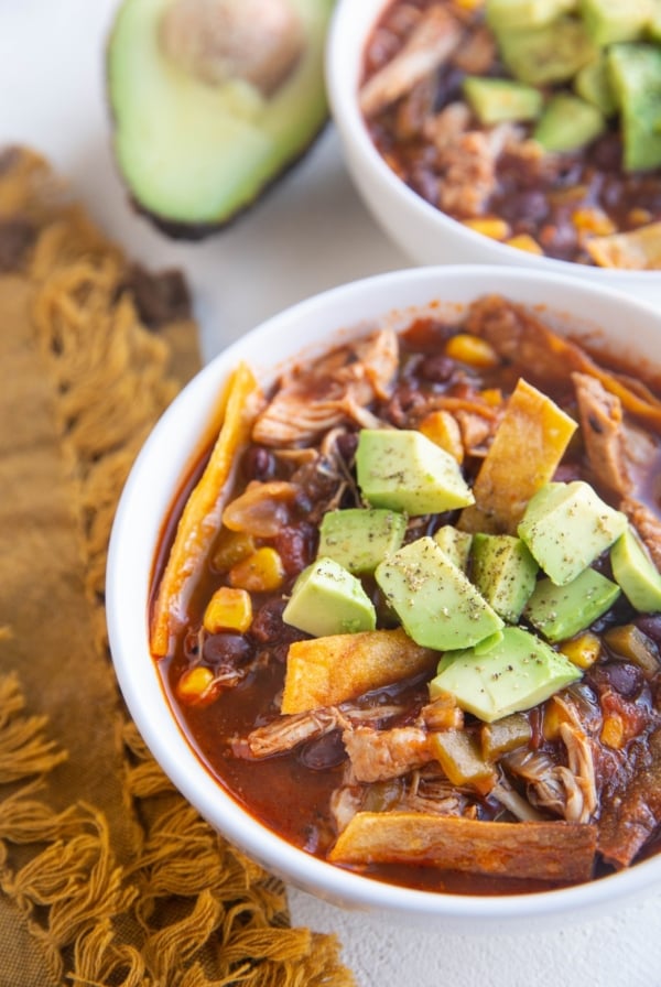 Two white bowls of chicken tortilla soup with avocado on top and a golden napkin to the side.